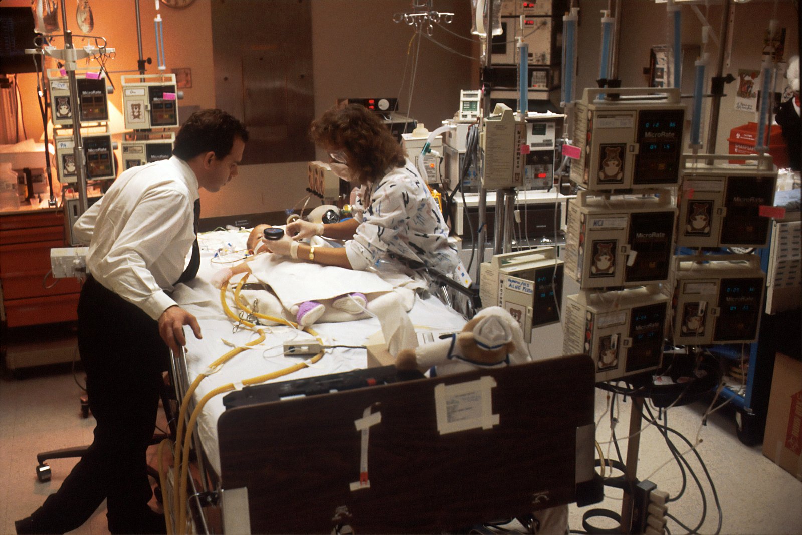 Two healthcare professionals in a clinical setting examine a patient on a bed surrounded by modern medical monitoring equipment and digital displays.